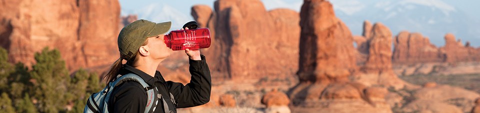 A woman drinks water out of a red bottle. Behind her is a dramatic scene of red rocks.