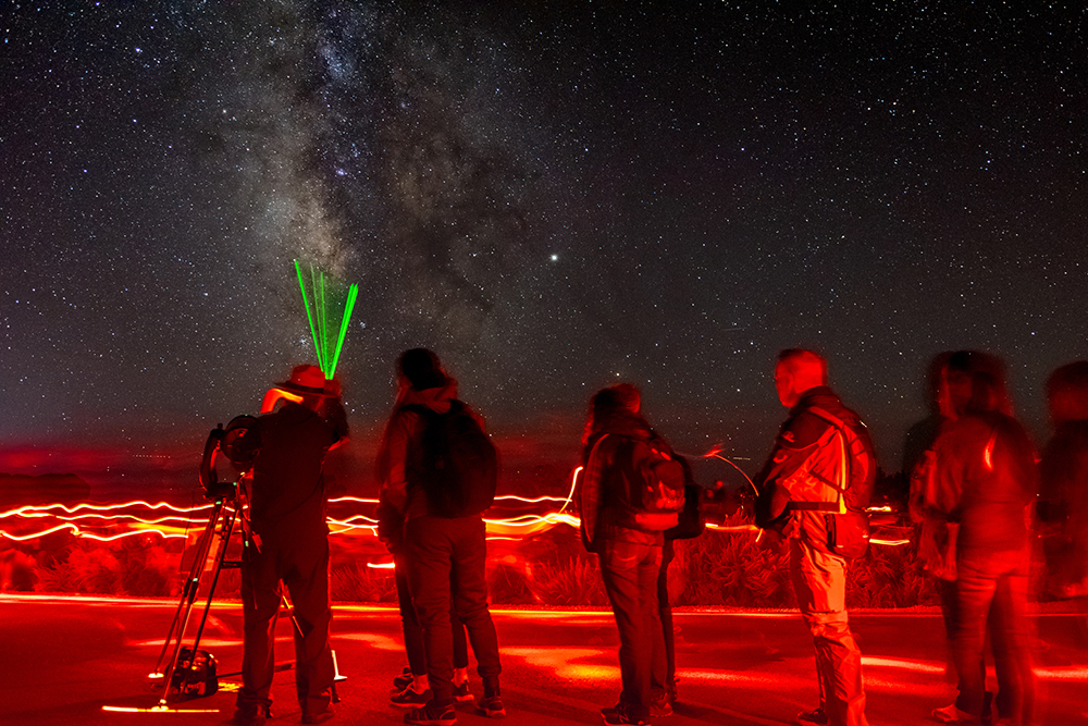 a ranger at a telescope uses a green laser pointer to show something in the starry night sky to a line of people.