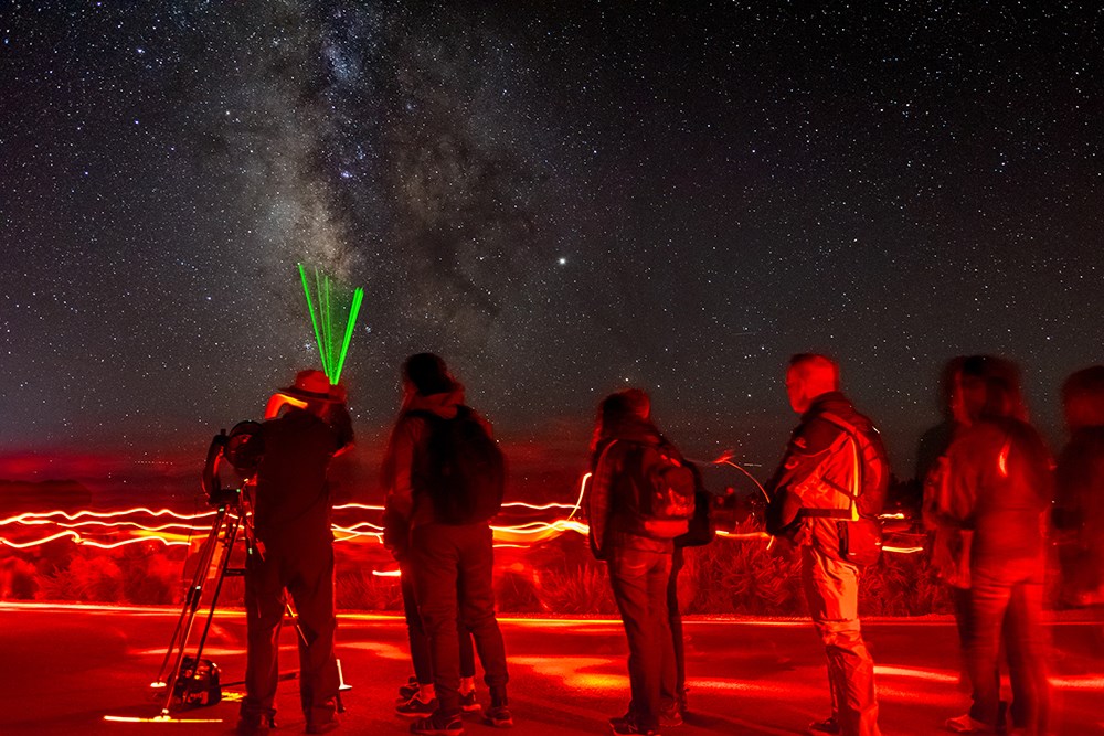 a ranger at a telescope uses a green laser pointer to show something in the starry night sky to a line of people.