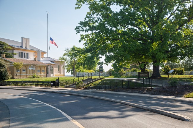 Stairs and a ramp lead from a road up to a sidewalk towards a two-story building and a flag flying at half-mast.