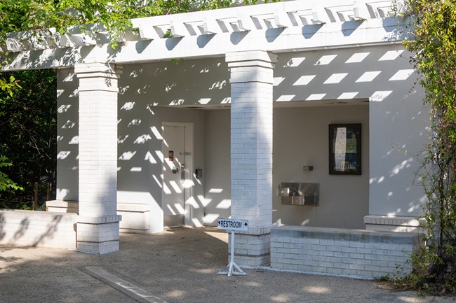 A one-story white brick building with two pillars, two doors to restrooms, and a drinking fountain.