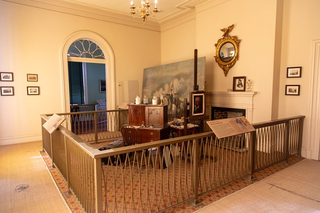 A room with tall ceilings and art supplies next to a large painting cordoned off by railings with information panels.