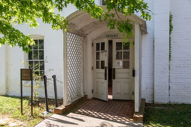 A brick path leads to a white brick building with double doors slightly ajar.