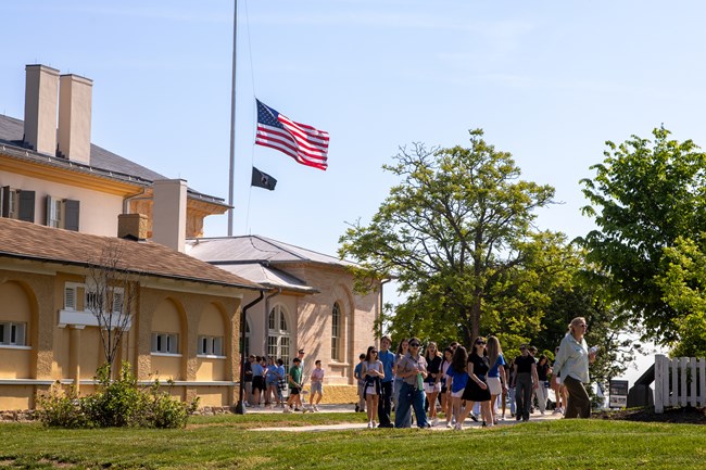An American flag at half mast waves over a two-story building with children walking on a sidewalk.