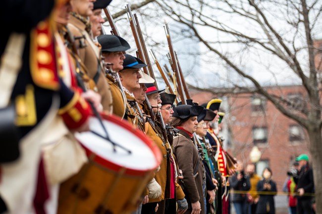 Row of people in 1770s clothing, holding muskets pointing up.