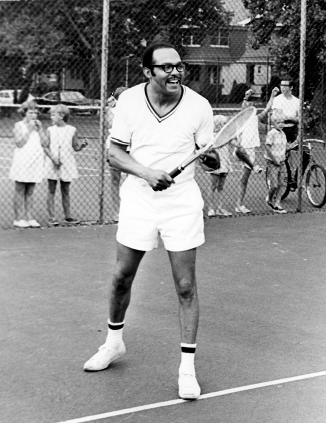 A Black man in glasses and a white outfit smiles and holds a racket on a tennis court as onlookers watch behind a fence.