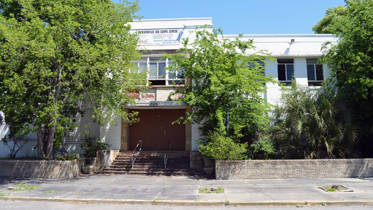 Exterior of the Jacksonville Jewish Center. Steps lead up to the building's wide front door. Trees are growing in front of the white and gray building in an area defined by a low garden wall.