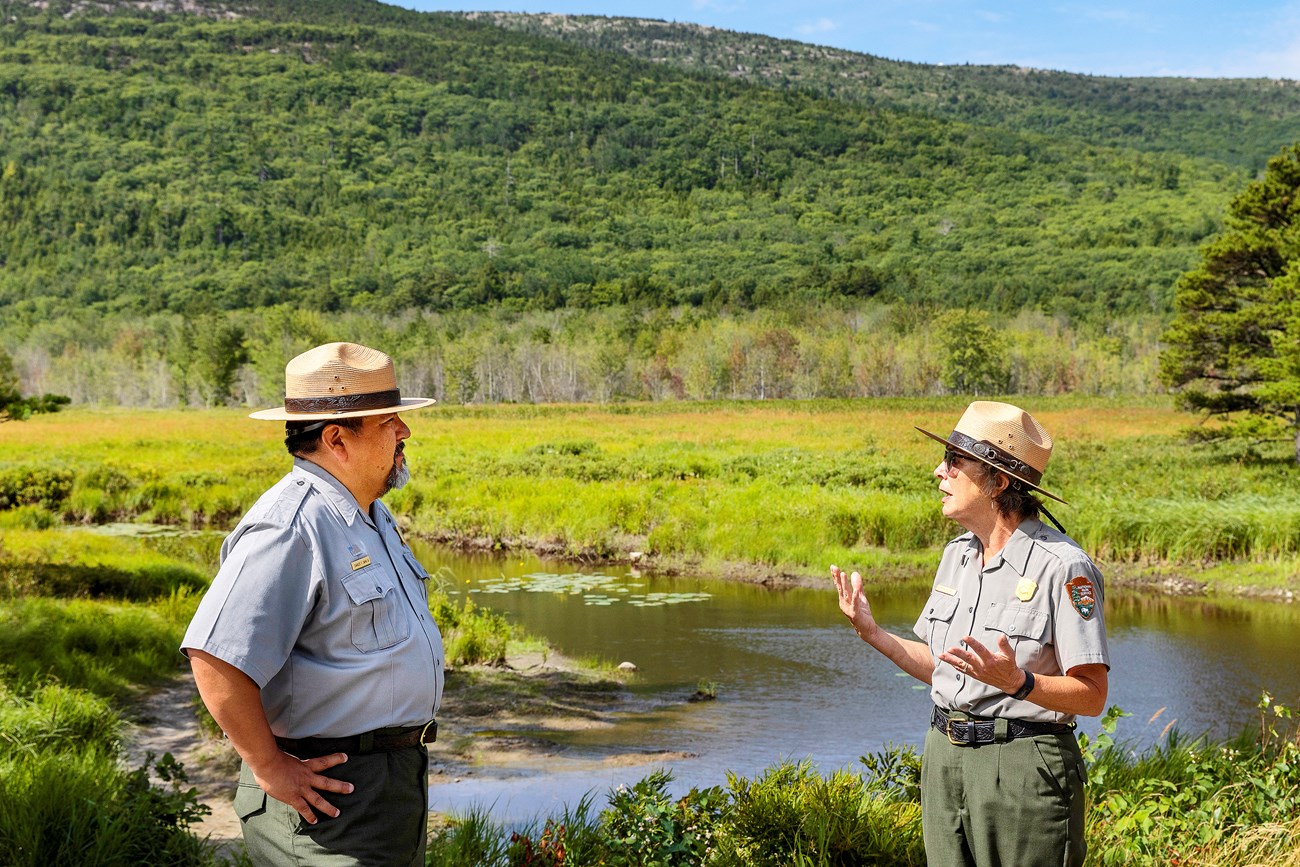 Two people in full NPS uniform. Director Sams, on the left, listens as Cole-Will talks and gestures, with the meadow and mountains behind them.