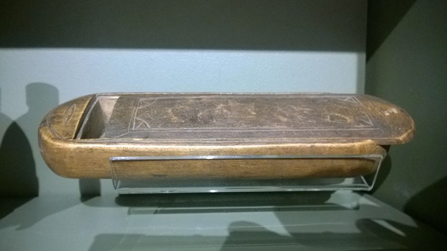 A small wooden rectangular box, with rounded ends and a simple carved ornamental design, sits behind glass in a museum display.  The sliding cover is partially open, showing the inside of the case, which is large enough to hold a shaving razor.