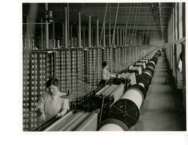 Black and white photo of a large room with two women working on machines. There are large structures with many bobbins of thread, each thread feeds into a machine that is producing a large roll of threads.