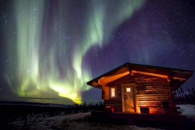 A night scene in Alaska.  In the foreground a small log cabin is illuminated with an artificial light.  To the left, in the sky, is the glow of light green curtain-like streaks of the aurora borealis.  Below is a river valley with trees and snow.