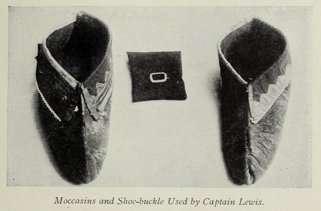 An old black and white photograph of two simple, one-piece leather moccasins, with a small shoe buckle positioned in between.