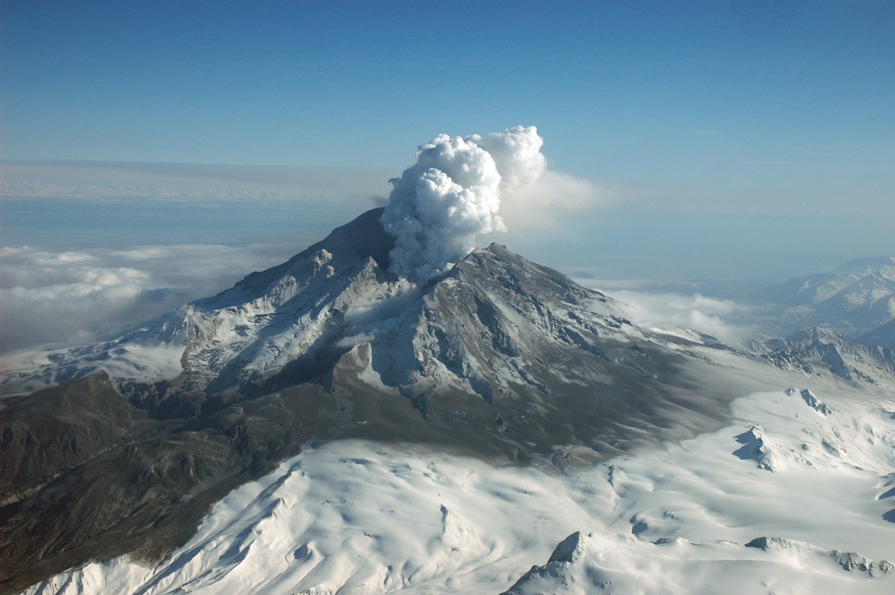 Photo of an erupting volcanic mountain with a steam cloud above the summit crater and snow on its lower slopes