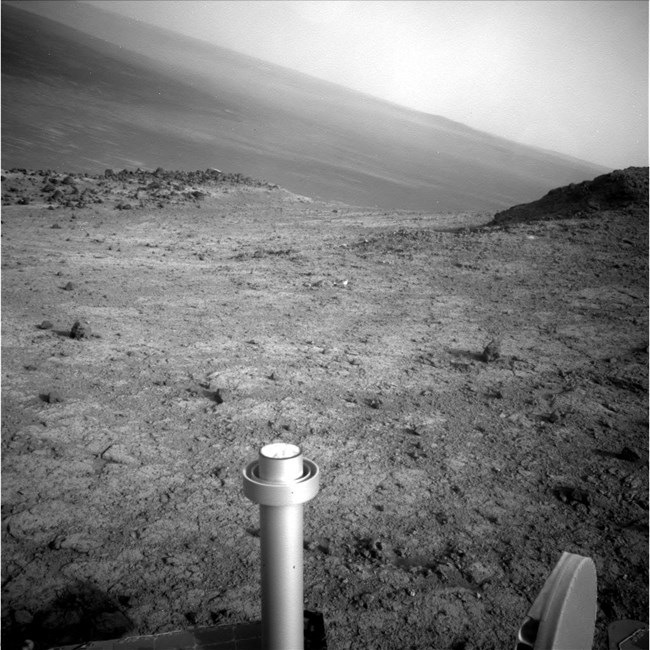 A black and white image from MER Opportunity.  Part of the rover is seen in the foreground.  The photo shows a rocky surface with a high ridge to the right.