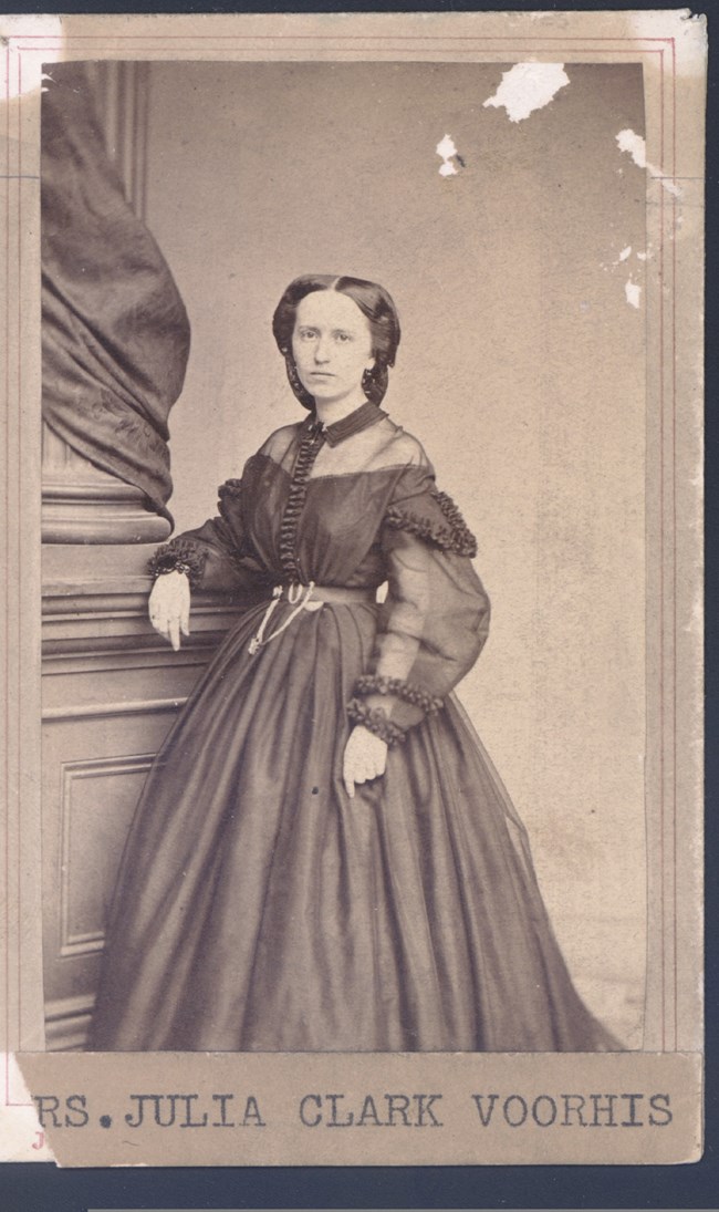 An old black and white photo shows Julia Clark Voorhis standing, leaning against a large column.  She is wearing a dark-colored dress of the period.