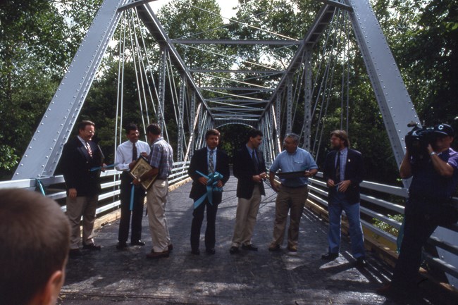 Seven men stand in a line across the bridge, chatting and holding plaques and cut ribbon with a TV cameraman, right.