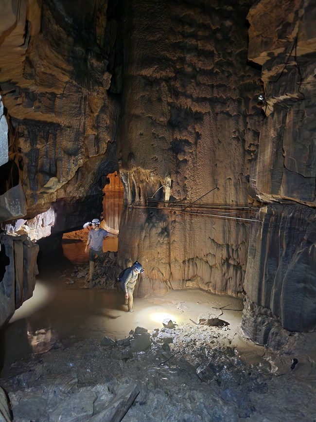 Two people with helmets and headlamps stand in shallow muddy water in a rocky cave room.