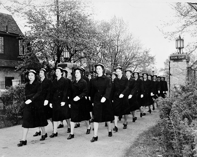 Four rows of uniformed women marching in line. They are wearing dark, dress coats that go down past the knee and their WAVE caps.