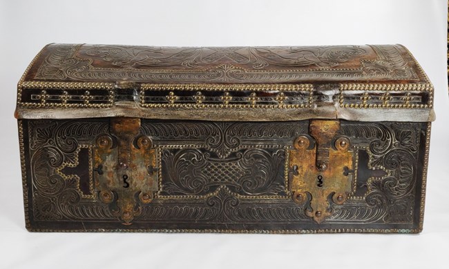 A dark rectangular trunk, with ornate designs in leather.  Two large metal latches are on the front.