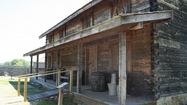 A rough-hewn cabin with a covered porch is in the foreground.  Behind it is the fort’s wooden palisade with a corner guard house.