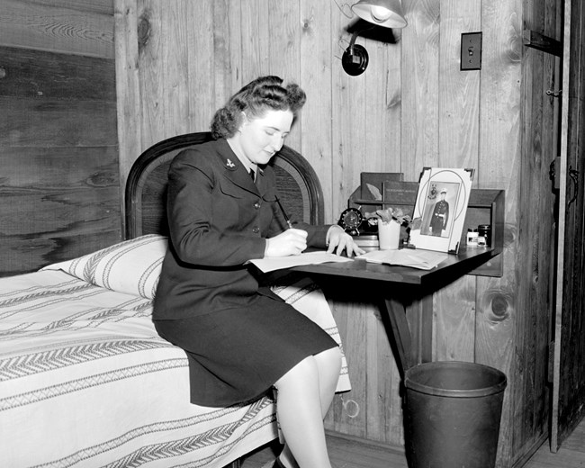 Woman wearing dark, uniform dress writes a letter on her bedside table while sitting on the edge of her bed.