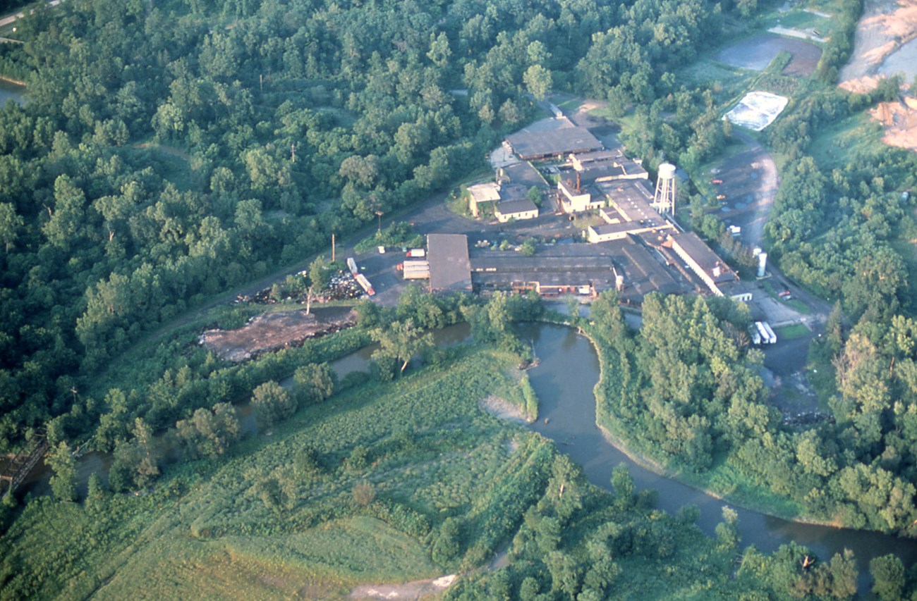 A bird’s-eye view of an industrial complex along a river bend, surrounded by greenery.