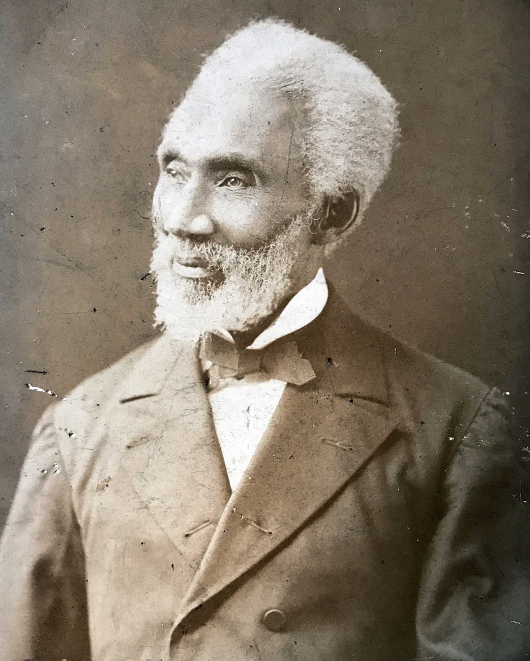 Bust-length photographic portrait of a grey or white-haired Black man with a beard looking towards his right, wearing a dark suit, light shirt, and a dark-colored bow tie.