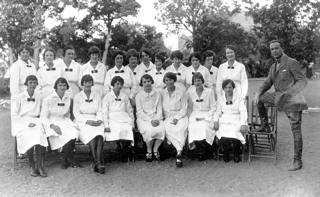 Fred Harvey General Manager Victor Patrosso, right, posing with group of 20 El Tovar Harvey Girls arranged in two rows, in in all white evening uniforms.