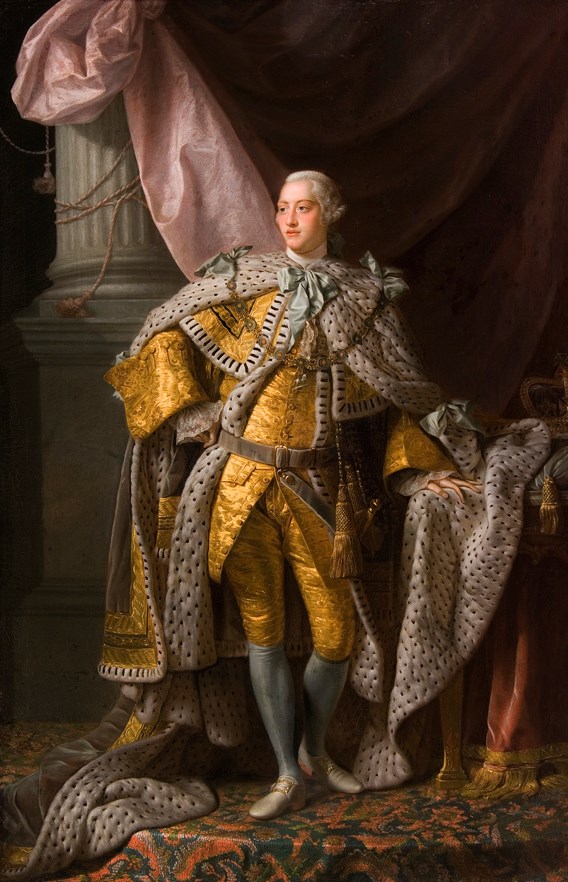 an adult white man with white powdered hair. He is wearing a gold outfit, resplendent with white royal robes. He is adorned with light blue bows and jewels. The background is lavish, with a pillar, curtain, and patterned rug. The royal crown sits to the l