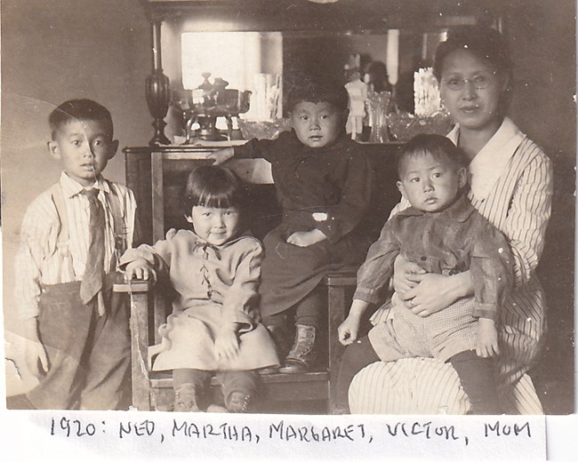 Family portrait. A young boy stands next to a chair that two young girls are sitting in. The mother sits next to them with another young boy in her lap.