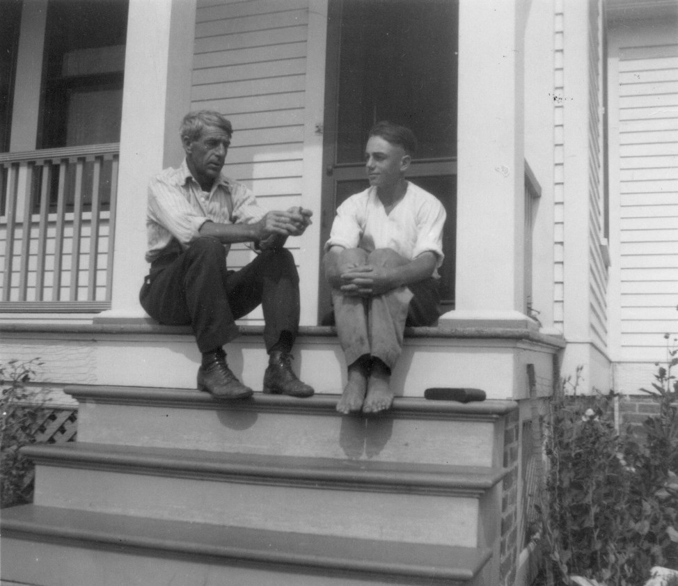 A father and teenage son sit on the front steps of a white house barefoot.