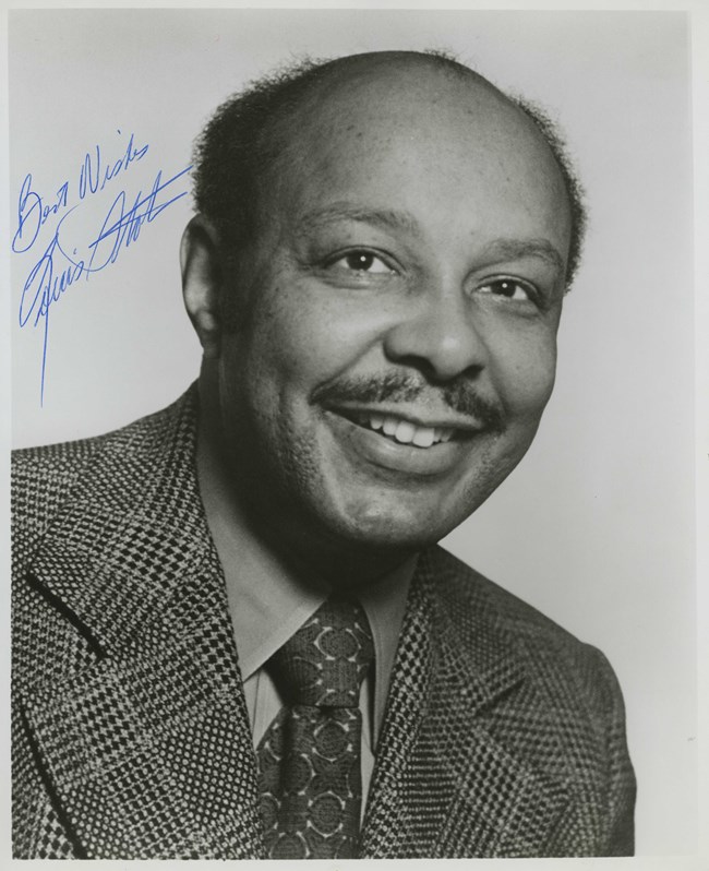 A studio portrait of a smiling Black man with a moustache wearing a patterned suit and tie; a blue autograph in the upper left corner.