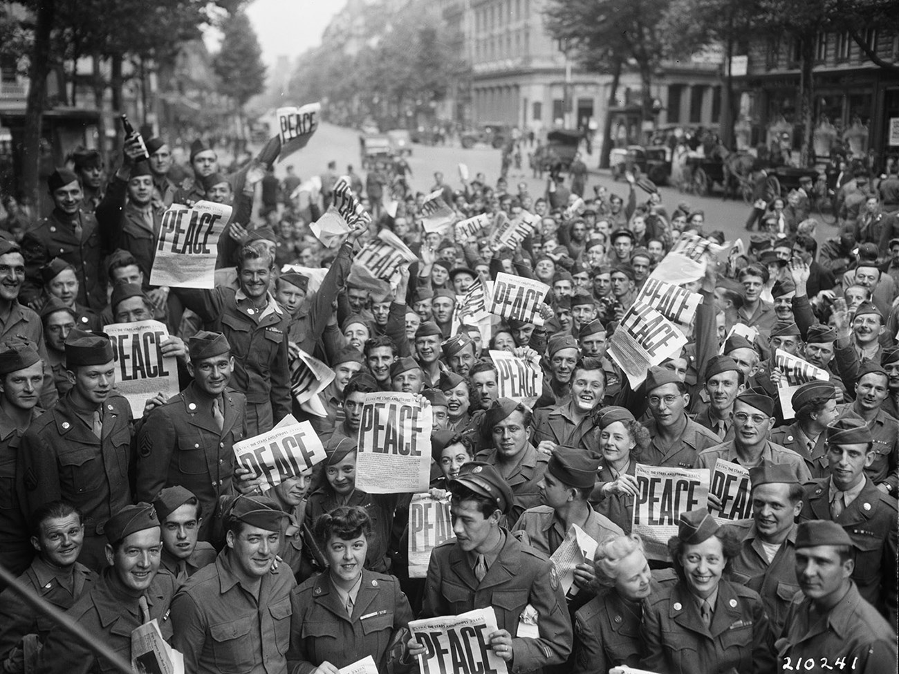 Many soldiers in uniform crowd a street holding newspapers that announce "Peace."