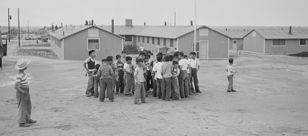 A group of young boys congregate on a dirt field in front of a large complex of buildings.