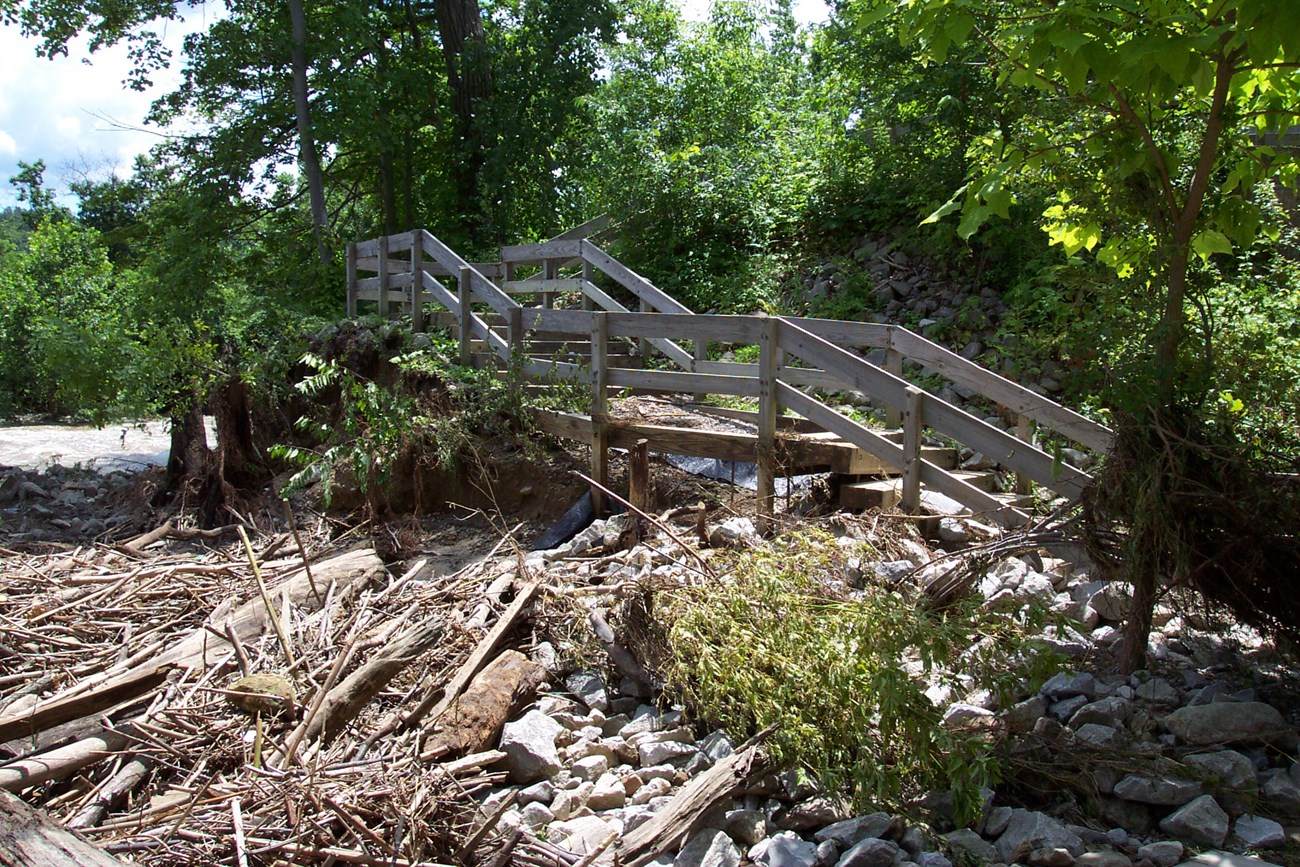 Beside a river, the floor of a wooden staircase is missing. Below is a jumbled pile of sticks and rocks.