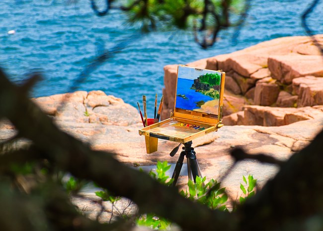 Through tree branches along a rocky coastline, an unattended easel holds a landscape painting in progress