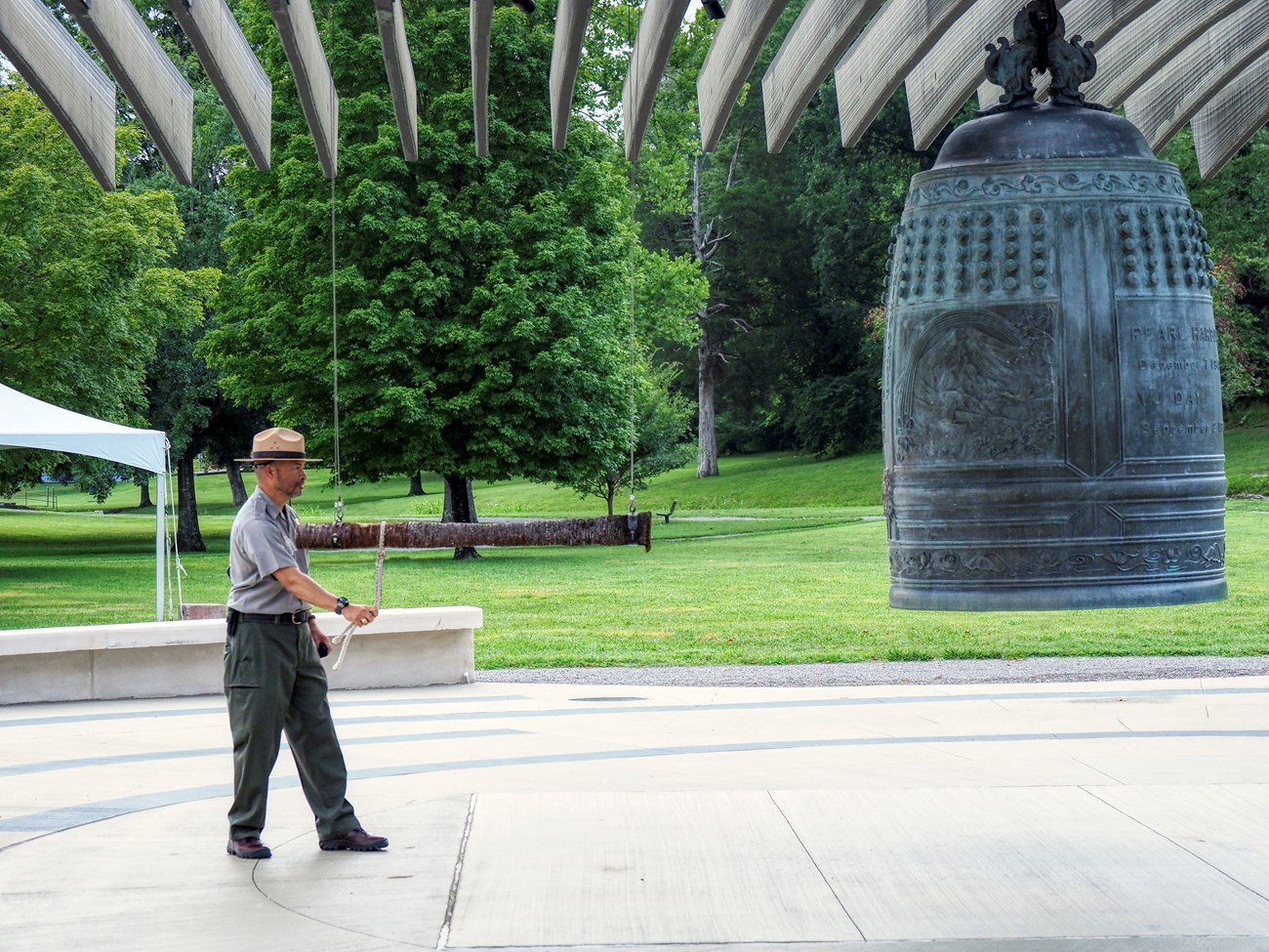 A uniformed park ranger holds a large horizontal bar about to ring a seven foot tall bronze, decorative commemoration bell in a park courtyard.