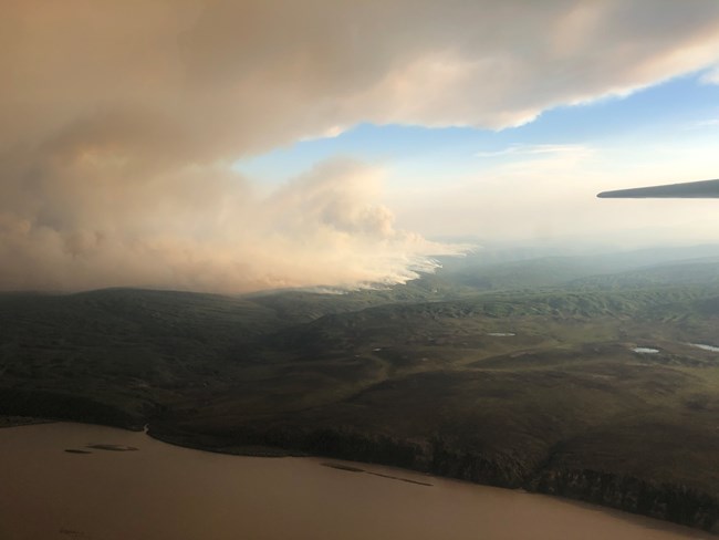 Image taken from aircraft showing billowing smoke from a forested area in the distance behind a large water body.