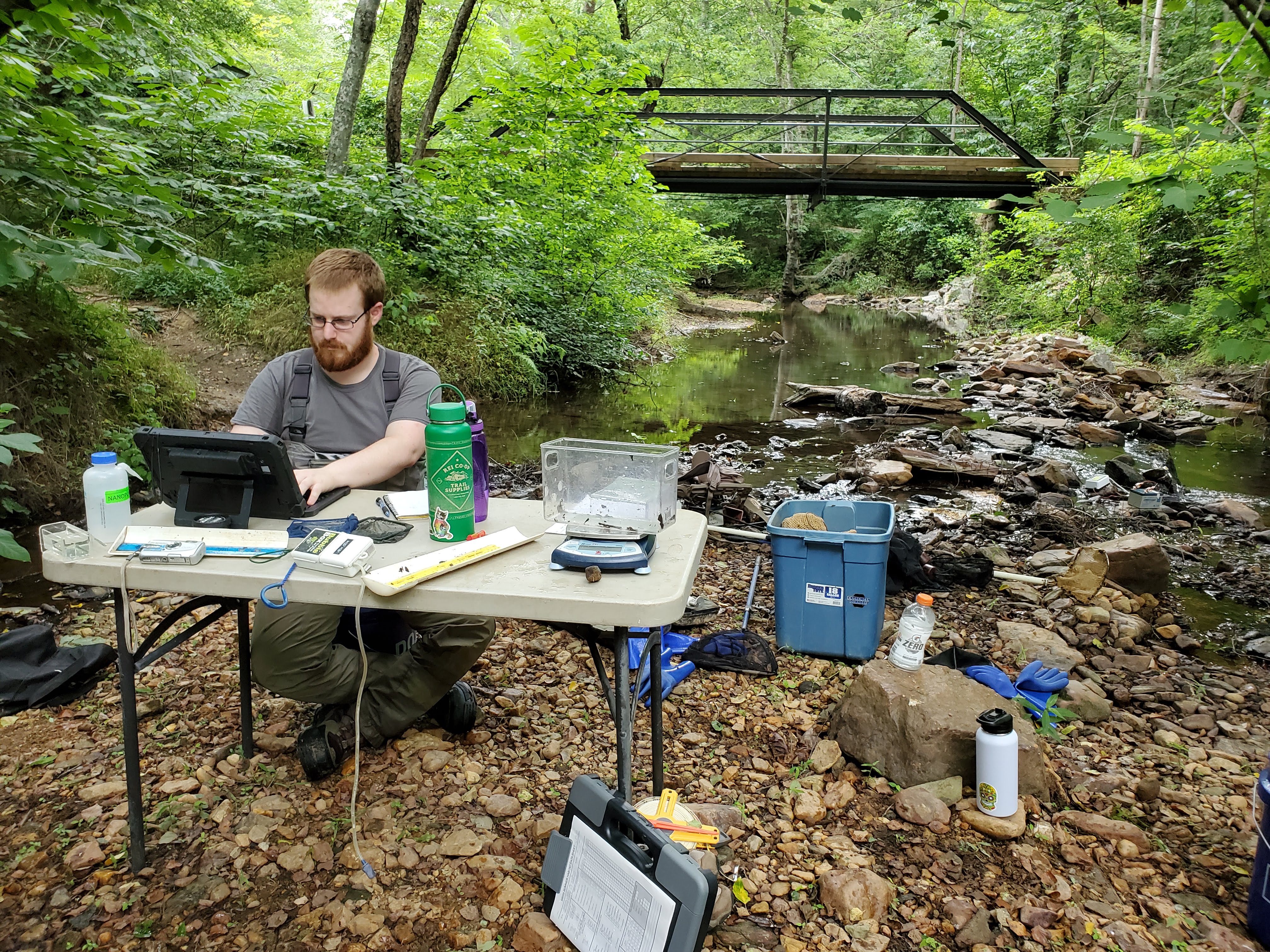 A scientist sits at a table on a stream bank, entering data into a computer