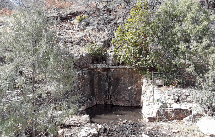 Square pool confined on three sides by rock walls on the edge of a drainage, with scattered trees on the slope around the pool and in the drainage.