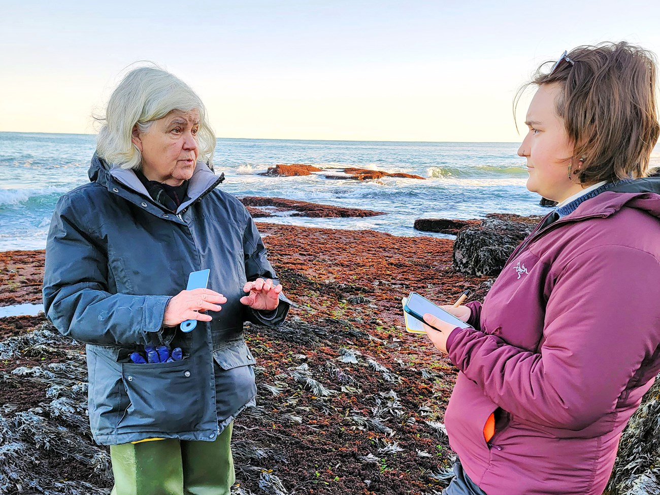 Older woman in green hip boots speaking to a younger woman holding a pad, pen, and cellphone. They're outside on a rocky, algae-covered stretch of coastline with the ocean behind them.