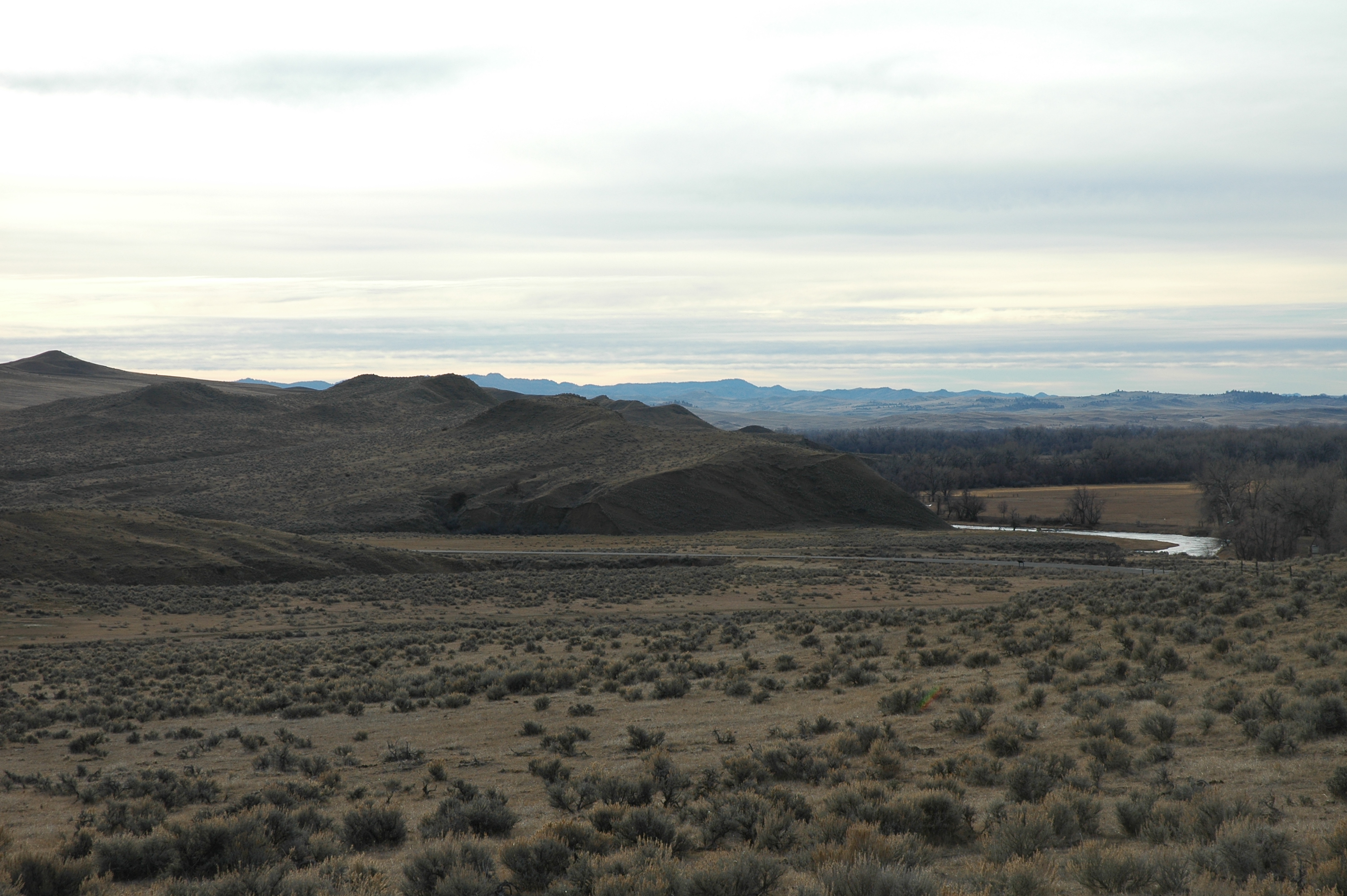 A large open prairie near several low gray hills and a small creek in the left-hand side of the image foreground. A distant chain of mountains are visible in the background. A small forest can be seen in between.