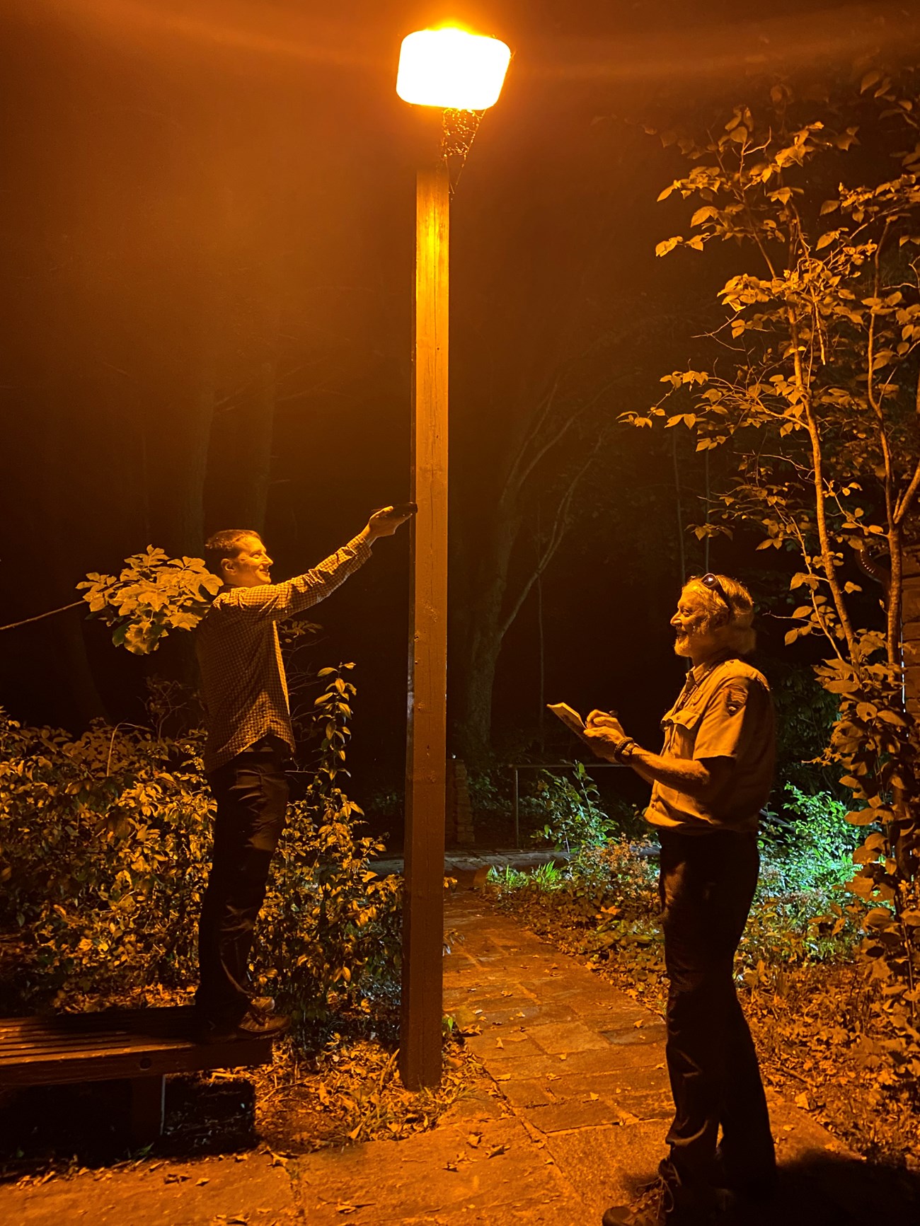 Two people stand below a light that is emitting an amber glow. One holds up an electronic measurement device as the other takes notes.