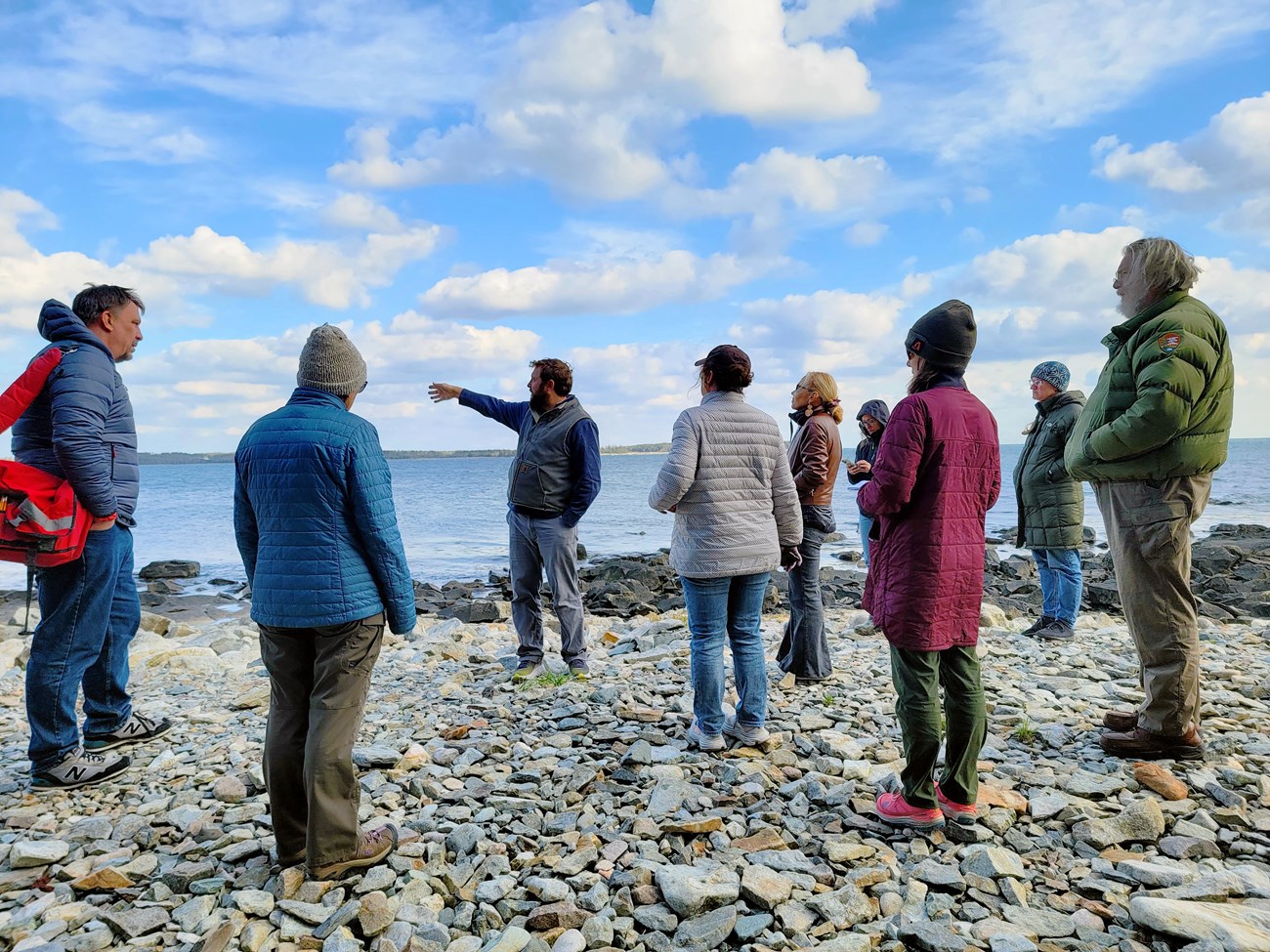 Group of people in jackets and hats stand along a rocky shore, their gaze following the gesturing arm of a man standing in the center.