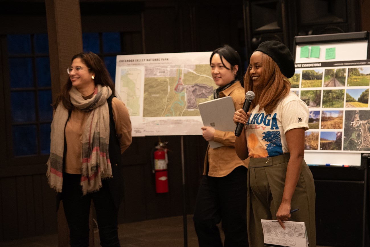 Three women stand in front of posters with maps and photos. One holds a microphone.