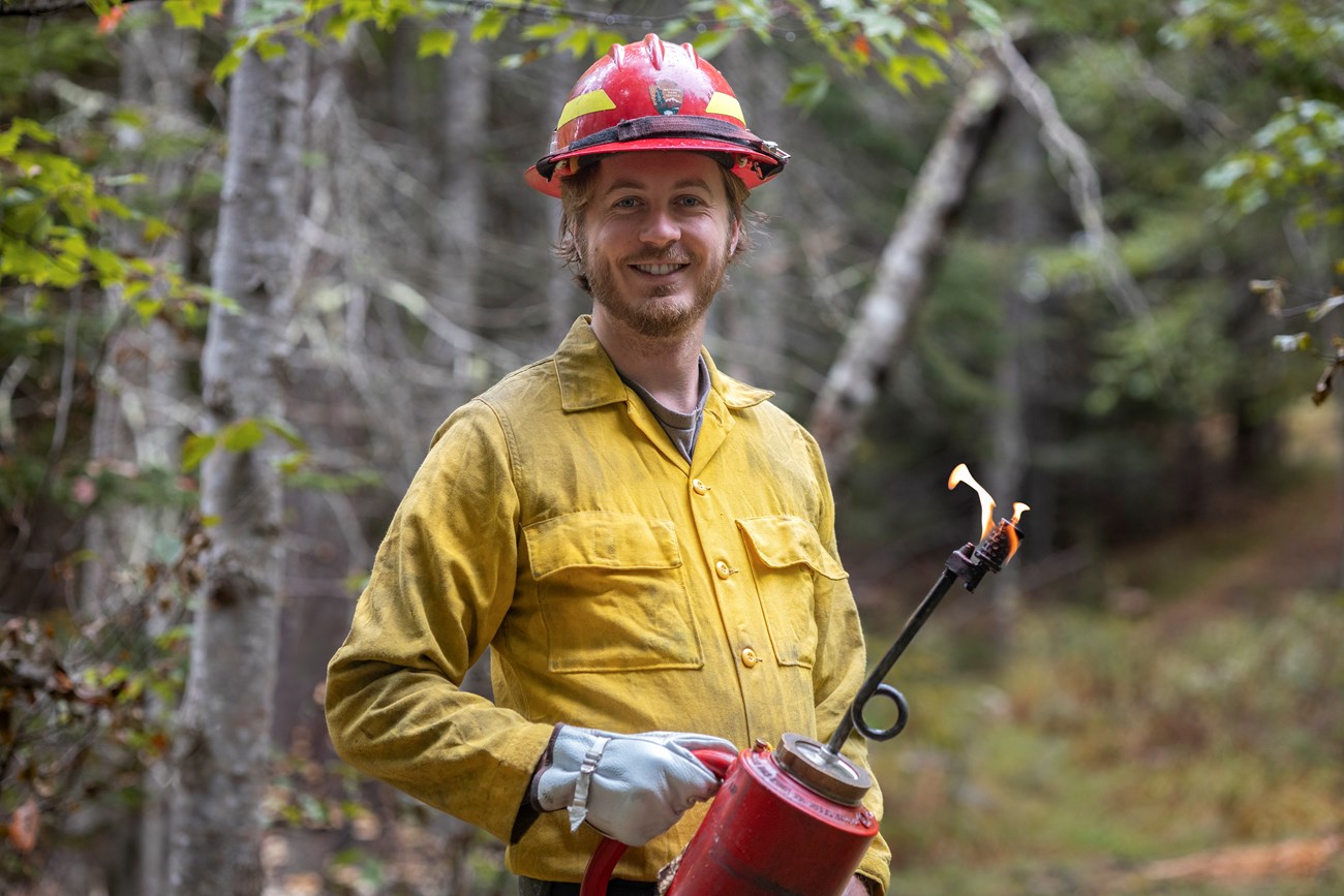 Aidan Gordon, Wildland Fire Apprentice, holds a flaming drip torch in a forested environment.