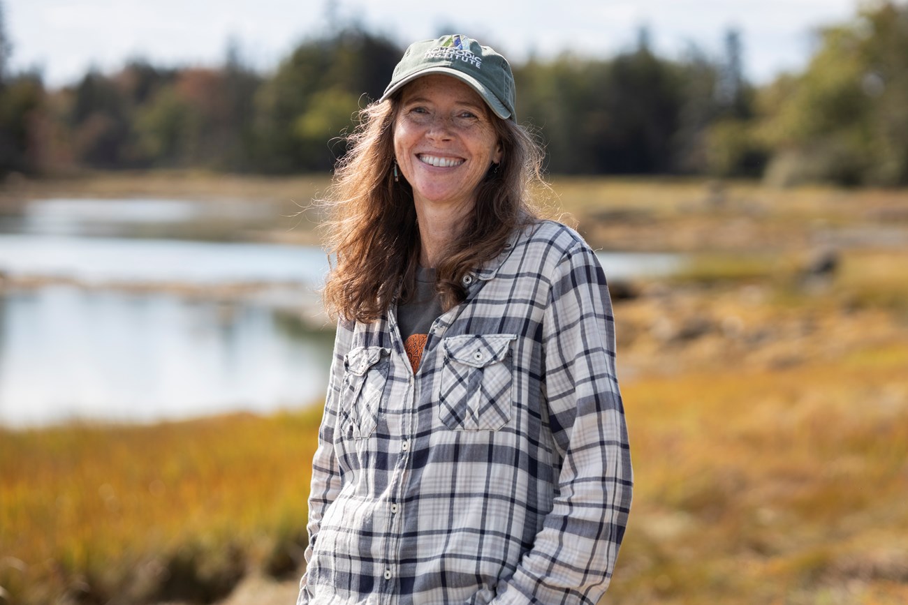 Woman wearing a hat stands in a wetland environment while smiling with the wind blowing her hair to the side.
