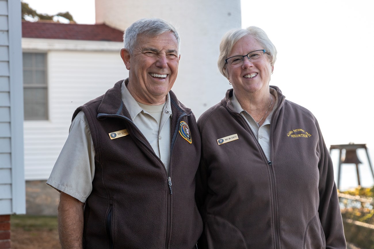 A husband and wife wearing volunteer uniforms smile in front of a lighthouse.