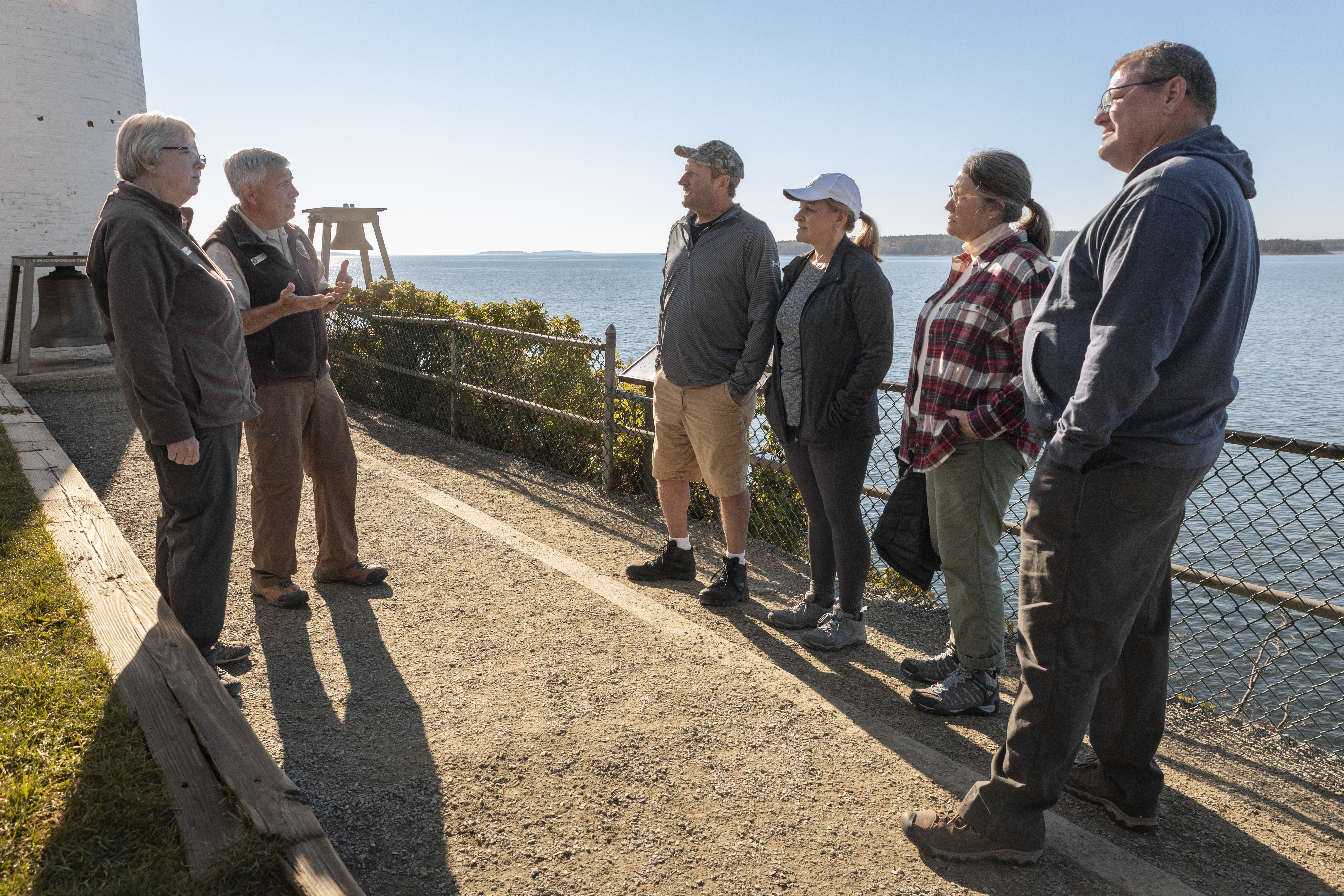Married volunteers talk to four visitors in front of a lighthouse.