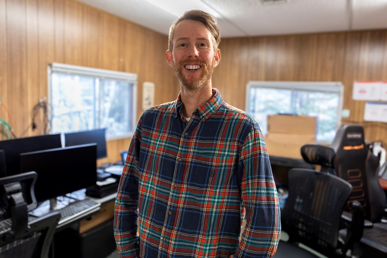 A man wearing a plaid shirt stands in a small office space filled with computers.
